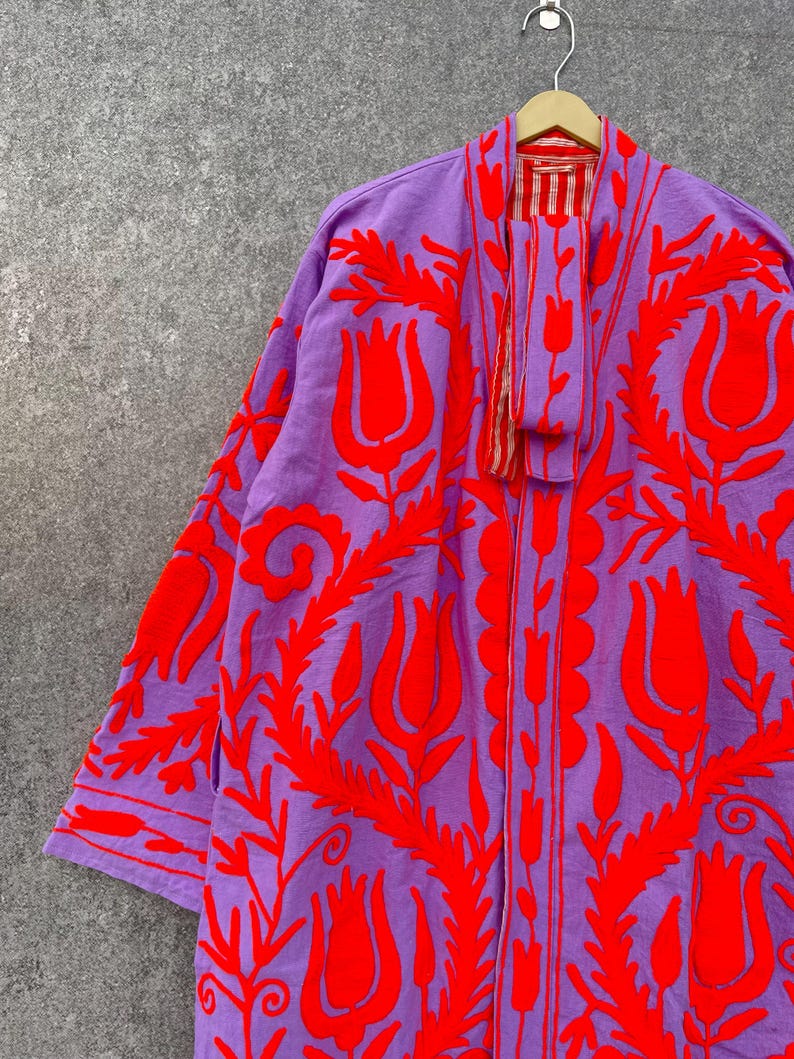 Puede incluir: Una larga t&uacute;nica morada con dise&ntilde;os florales bordados en rojo. La t&uacute;nica tiene un forro a rayas rojo y blanco visible en el cuello.