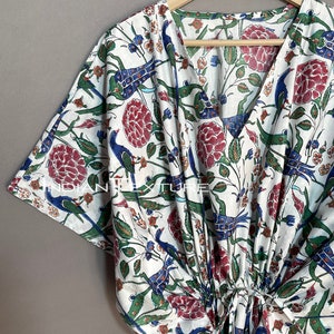 May include: White, V-neck kaftan with a colorful floral and bird print. The design features red flowers, blue birds, and green leaves. The kaftan is hanging on a wooden hanger.