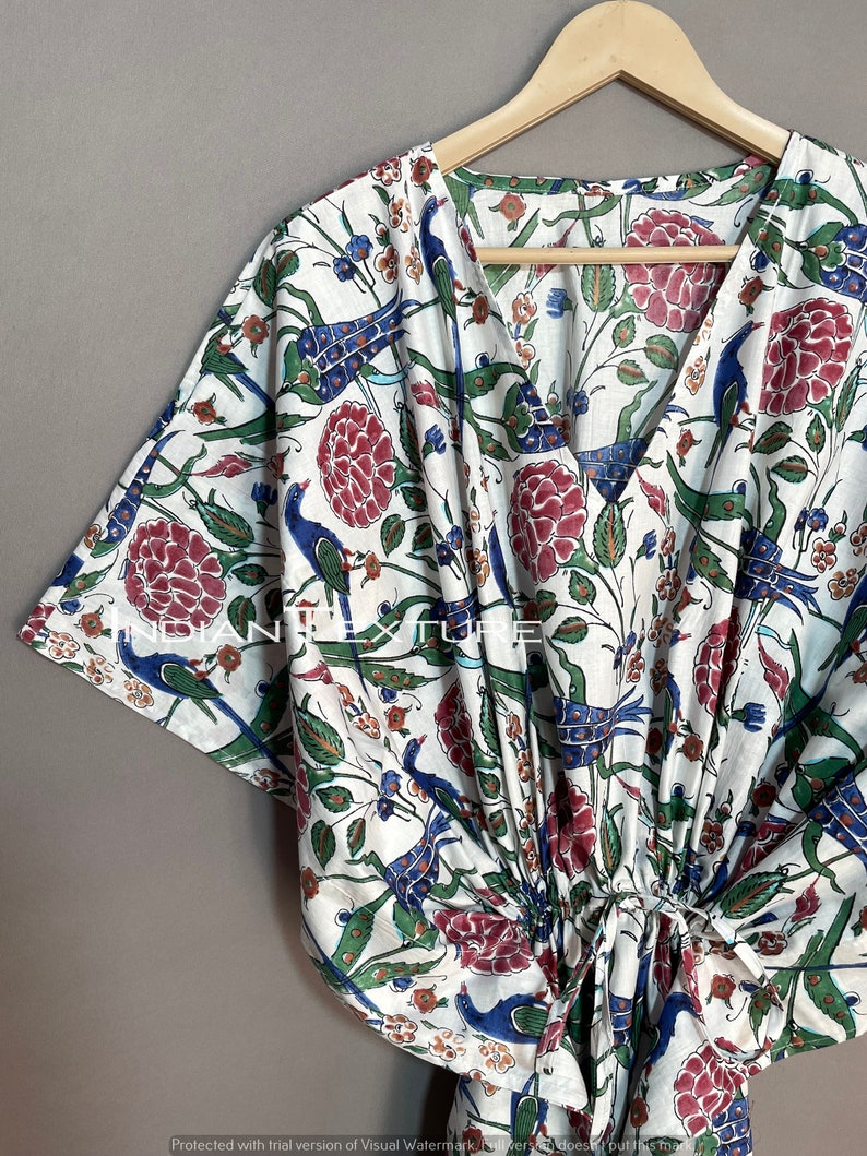 May include: White, V-neck kaftan with a colorful floral and bird print. The design features red flowers, blue birds, and green leaves. The kaftan is hanging on a wooden hanger.