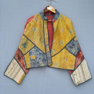 Peut inclure: Une veste courte présentant un motif patchwork composé de divers tissus en jaune, bleu et rouge. Le vêtement possède des manches longues, un col montant et un liseré gris sur les bords. Une petite étiquette blanche avec le texte Indian Texture est sur le cintre.