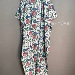 May include: A white caftan with a vibrant floral and bird pattern. The design features red flowers, blue birds, and green foliage. The caftan is hanging on a wooden hanger against a neutral background. The word "EXTURE" is visible.