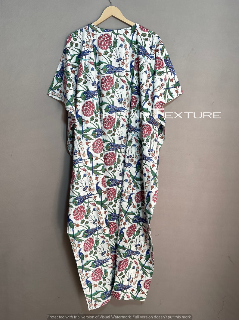 May include: A white caftan with a vibrant floral and bird pattern. The design features red flowers, blue birds, and green foliage. The caftan is hanging on a wooden hanger against a neutral background. The word "EXTURE" is visible.