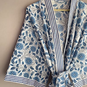 May include: A white and blue floral print robe with a blue and white striped trim. The robe has a tie belt and a pocket on the left side. The text "INDIAN TEXTURE" is written on the wooden hanger.