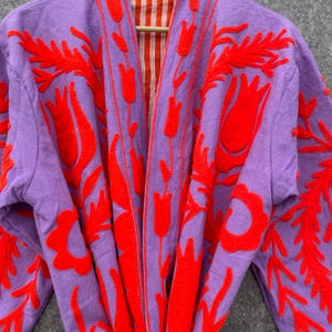 Puede incluir: Una bata morada con un dise&ntilde;o de bordado floral rojo. La bata tiene un forro a rayas.