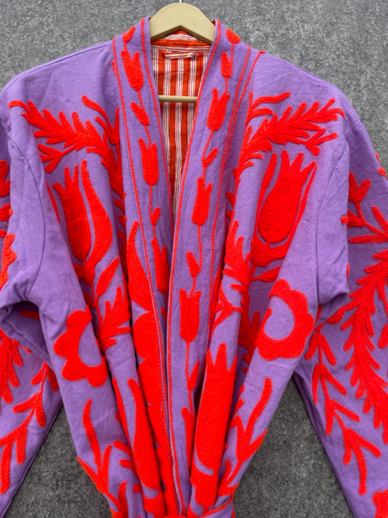 Puede incluir: Una bata morada con un dise&ntilde;o de bordado floral rojo. La bata tiene un forro a rayas.