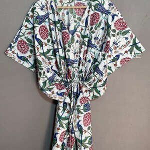 May include: White caftan with a V-neck and a floral and bird pattern in red, blue, and green. The caftan is hanging on a wooden hanger. The fabric is lightweight and appears to be cotton.