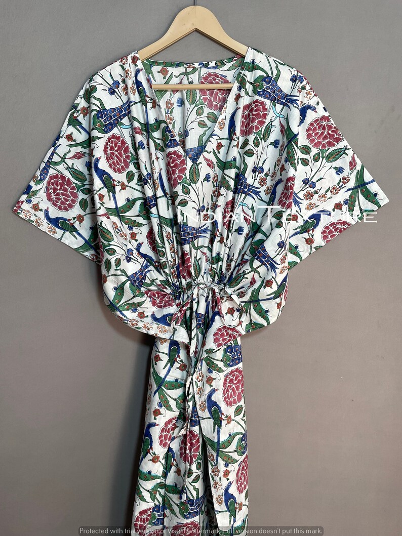 May include: White caftan with a V-neck and a floral and bird pattern in red, blue, and green. The caftan is hanging on a wooden hanger. The fabric is lightweight and appears to be cotton.