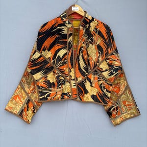 Puede incluir: Una chaqueta corta con un patrón vibrante de motivos florales en negro, naranja y dorado. La prenda está confeccionada con costuras acolchadas visibles y acabada con ribetes de tela marrón a lo largo de los bordes, el cuello y las costuras.