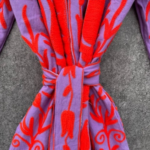 Puede incluir: Una bata morada con un dise&ntilde;o de bordado floral rojo. La bata tiene un cintur&oacute;n de lazo y est&aacute; hecha de una tela suave y c&oacute;moda.