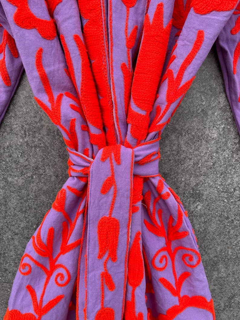 Puede incluir: Una bata morada con un dise&ntilde;o de bordado floral rojo. La bata tiene un cintur&oacute;n de lazo y est&aacute; hecha de una tela suave y c&oacute;moda.