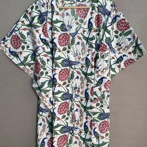 May include: White caftan with a V-neck and short sleeves, adorned with a floral and bird pattern in red, blue, and green. The fabric is lightweight, and the design includes a drawstring waist.