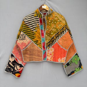 Puede incluir: Una chaqueta corta confeccionada con paneles de tela acolchada que presentan diversos patrones, incluyendo motivos florales, rayas blancas y negras y diseños geométricos en tonos naranja, amarillo, verde y negro, con ribetes gris claro.