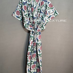 May include: A white, floral-patterned caftan with a V-neck and drawstring waist. The fabric features a print of blue birds, red flowers, and green leaves. The caftan is hanging on a wooden hanger.