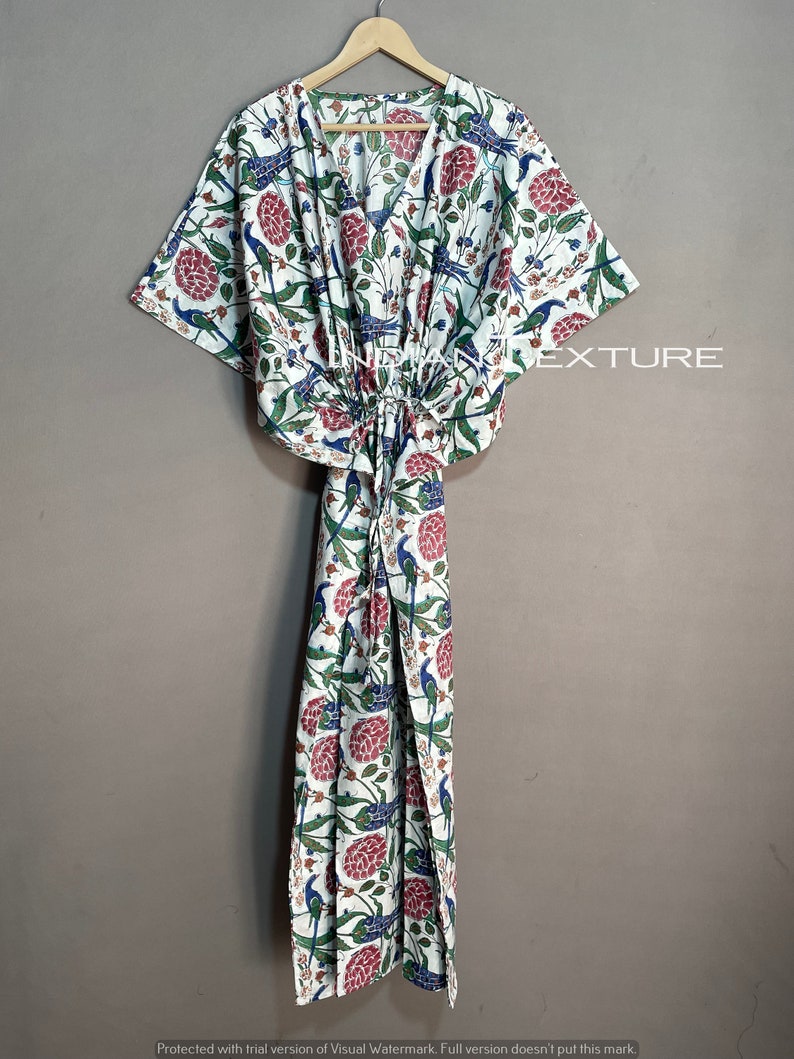 May include: A white, floral-patterned caftan with a V-neck and drawstring waist. The fabric features a print of blue birds, red flowers, and green leaves. The caftan is hanging on a wooden hanger.