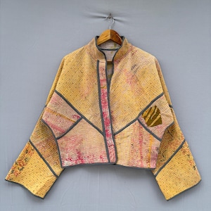 Op de afbeelding: Een kort jasje van gele en roze doorgestikte stof met zichtbare kantha-stiksels. Het jasje heeft een hoge kraag, wijde mouwen en donkergrijze biezen langs de randen, met een klein patch-detail op het voorste linkerzakgedeelte.