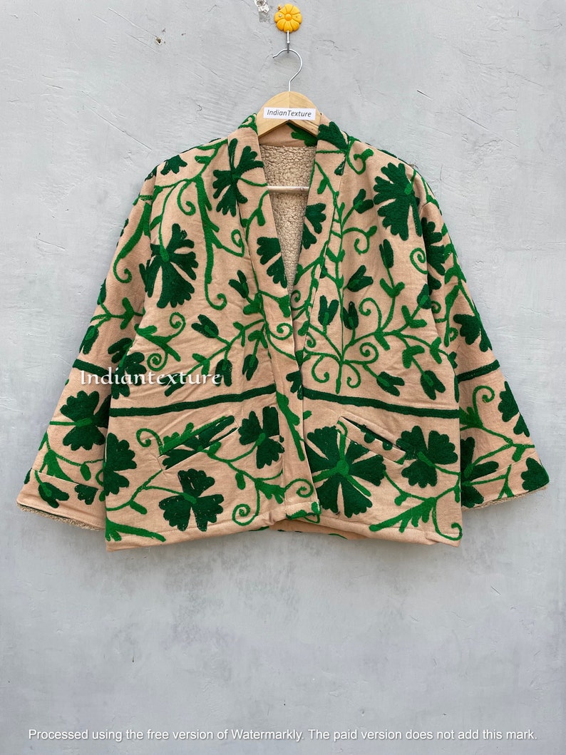 Op de afbeelding: Een beige geborduurde jas met een groen bloemenpatroon. De jas heeft een shawlkraag en lange mouwen. De jas is gevoerd met een zacht, pluche materiaal.