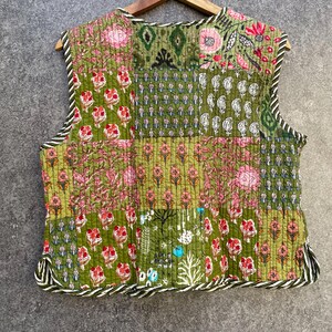 May include: A sleeveless quilted waistcoat with a patchwork design. The waistcoat is made of green fabric with floral patterns in pink, white, and blue. The waistcoat has a white and black striped trim around the edges.