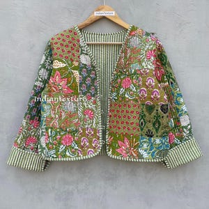 Op de afbeelding: Een groen gewatteerd jack met een bloemig patchwork-ontwerp. Het jack heeft een V-hals en een knoopsluiting. De mouwen zijn 3/4 lang en hebben een gestreepte afwerking.