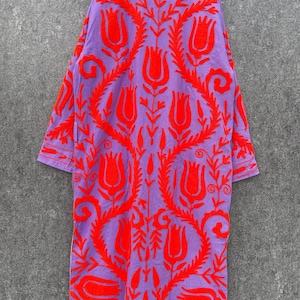 Puede incluir: Una larga t&uacute;nica morada con un patr&oacute;n de bordado floral rojo. La t&uacute;nica tiene cuello y mangas largas.