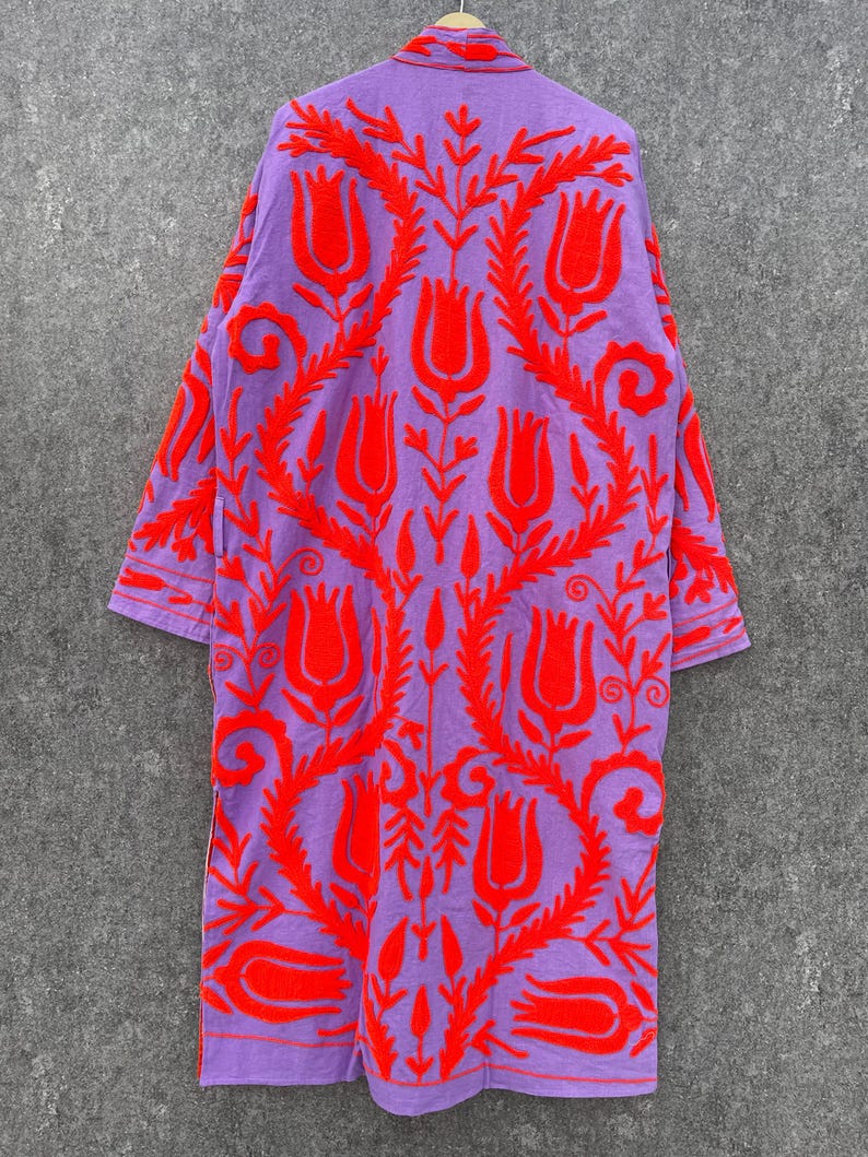 Puede incluir: Una larga t&uacute;nica morada con un patr&oacute;n de bordado floral rojo. La t&uacute;nica tiene cuello y mangas largas.