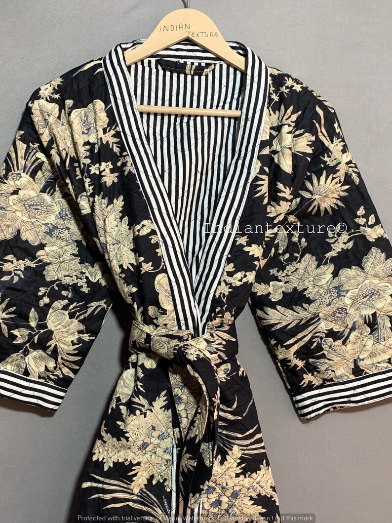 May include: A black quilted robe with a floral pattern in beige and blue. The robe has a striped white and black collar and cuffs. The robe is tied with a matching belt. The text "INDIAN TEXTURE" is visible on the hanger.