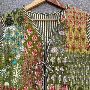 May include: A green quilted vest with a patchwork design featuring floral patterns in shades of pink, red, and blue. The vest has a V-neck and a tie closure at the front.
