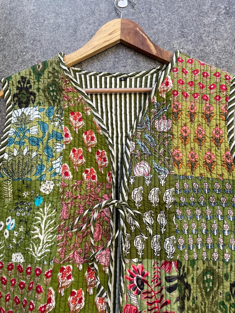 May include: A green quilted vest with a patchwork design featuring floral patterns in shades of pink, red, and blue. The vest has a V-neck and a tie closure at the front.