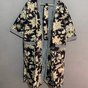 May include: A black floral print robe with a striped center panel and trim. The robe features a beige floral pattern on a black background. The sleeves and pockets are trimmed with white and black stripes. The robe is hanging on a wooden hanger.