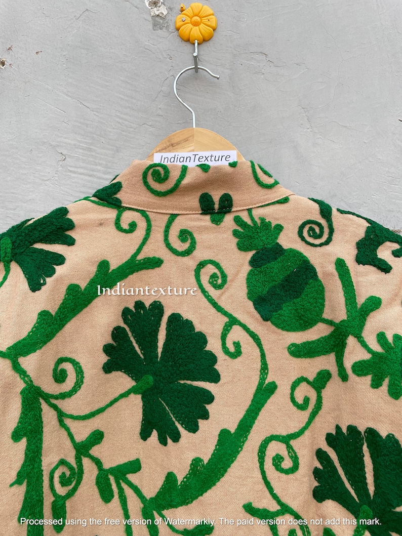 Op de afbeelding: Een beige katoenen jas met groene geborduurde bloemenmotieven. De jas heeft een kraag en een knoopsluiting. De borduursels omvatten spiralen, bladeren en bloemen.