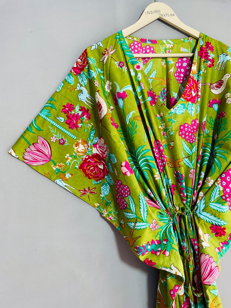 Peut inclure: Un kaftan en coton vert &agrave; imprim&eacute; floral avec des fleurs roses, blanches et bleues, des oiseaux et d'autres motifs. Le kaftan a un col en V et une ceinture &agrave; la taille. Le texte "INDIAN TEXTURE" est imprim&eacute; sur un cintre en bois.