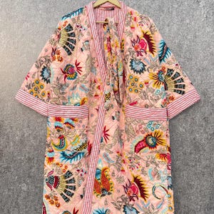 Könnte beinhalten: Eine rosa gesteppte Robe mit Blumendruck und gestreiftem Saum. Die Robe hat einen Bindegürtel und lange Ärmel.