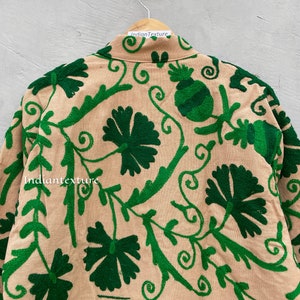 Op de afbeelding: Een beige katoenen jas met groene borduursels van bloemen en wijnstokken. De borduursels zijn gedetailleerd en ingewikkeld. De jas hangt aan een hanger.