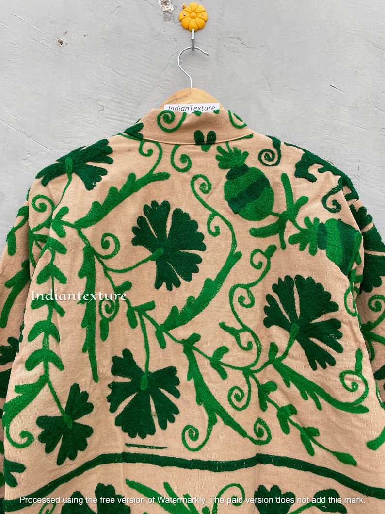 Op de afbeelding: Een beige katoenen jas met groene borduursels van bloemen en wijnstokken. De borduursels zijn gedetailleerd en ingewikkeld. De jas hangt aan een hanger.