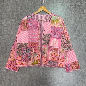Puede incluir: Una chaqueta acolchada rosa con un diseño de patchwork que presenta varios patrones florales y geométricos. La chaqueta tiene cierre de botones y mangas largas.
