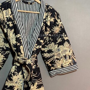 May include: A black quilted robe with a floral pattern in beige and blue. The robe has a striped white and black lining and trim. The robe is hanging on a wooden hanger. The text "INDIAN TEXTURE" is visible on the hanger.