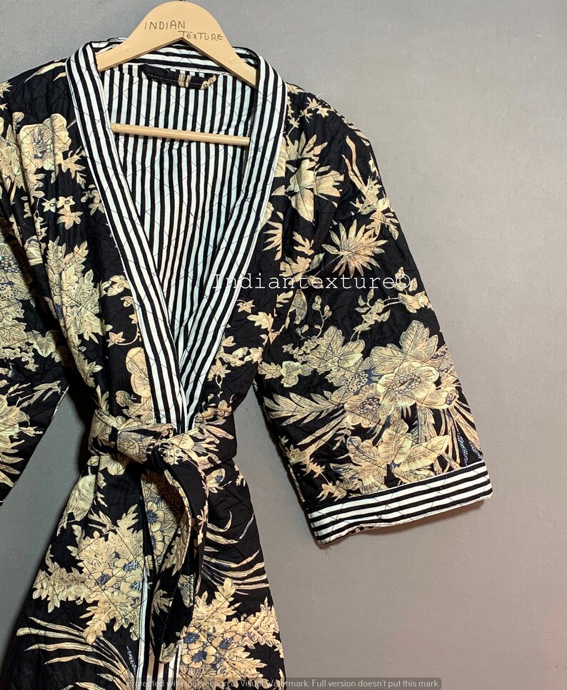 May include: A black quilted robe with a floral pattern in beige and blue. The robe has a striped white and black lining and trim. The robe is hanging on a wooden hanger. The text "INDIAN TEXTURE" is visible on the hanger.