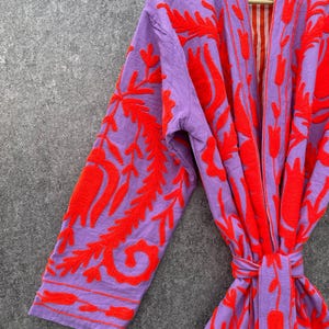 Puede incluir: Una bata morada con un patr&oacute;n de bordado floral rojo. La bata tiene un cintur&oacute;n de atar y un forro a rayas.