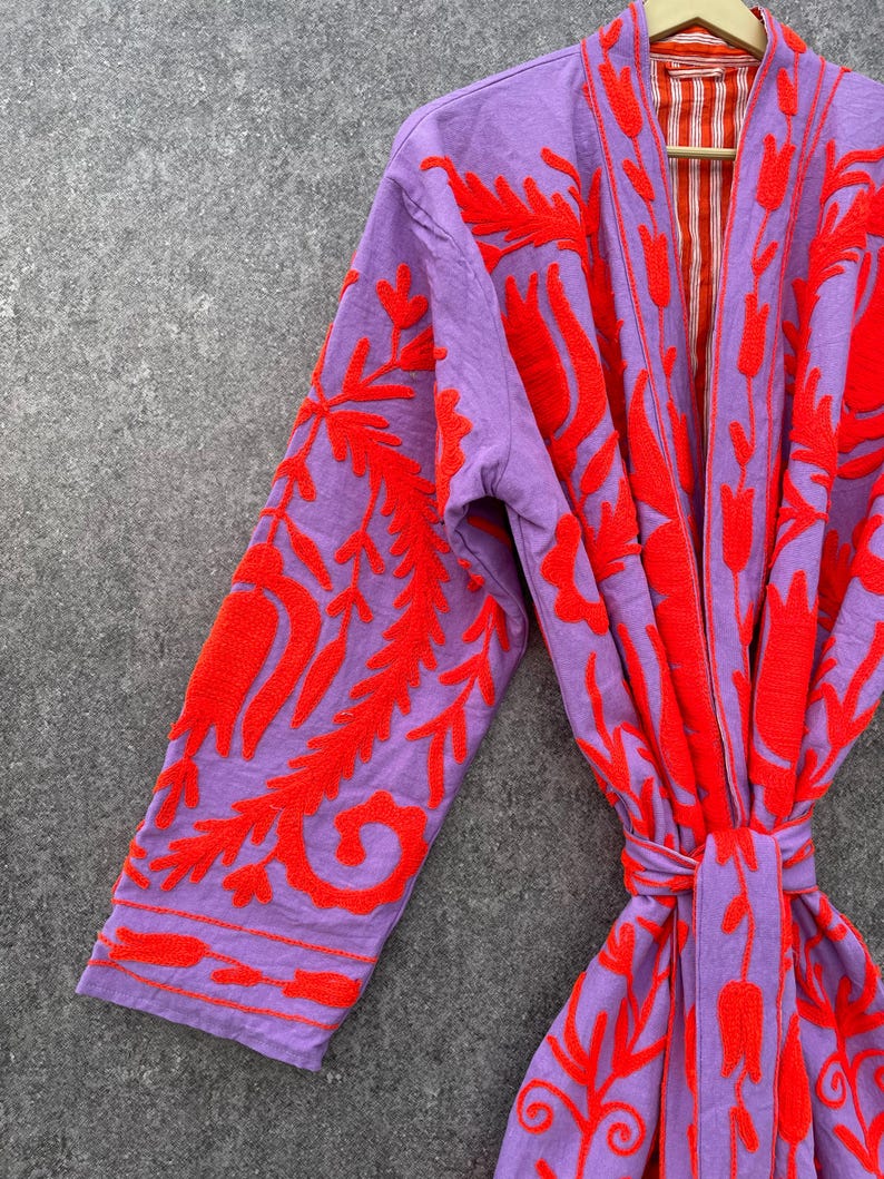 Puede incluir: Una bata morada con un patr&oacute;n de bordado floral rojo. La bata tiene un cintur&oacute;n de atar y un forro a rayas.