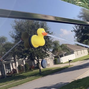 May include: A yellow rubber duck with a black base is attached to the back window of a black car. The duck is spraying a stream of water out of its beak.