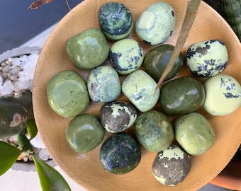 May include: A collection of green and white tumbled stones, also known as green aventurine, on a wooden surface. The stones are smooth and polished, with a variety of patterns and textures.