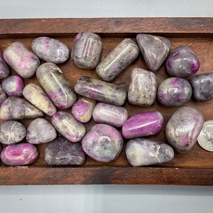 Könnte beinhalten: Eine Sammlung polierter rosa und grauer Steine in einer Holzkiste. Die Steine sind glatt und poliert und in einem zufälligen Muster angeordnet. Eine deutsche 1-Euro-Münze ist zur Größenbestimmung enthalten.