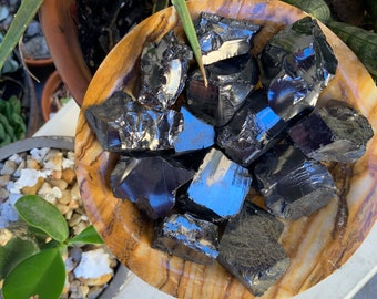 Könnte beinhalten: Eine Sammlung polierter schwarzer Obsidiansteine in einer dekorativen, runden, braun-beigen Schale. Die Steine haben eine reflektierende, glasartige Oberfläche. Die Schale steht vor einem Hintergrund aus Grünpflanzen.