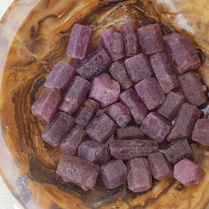 May include: A bowl filled with a collection of small, rough, purple ruby crystals. The crystals are various shapes and sizes, with some being more rectangular and others more rounded.