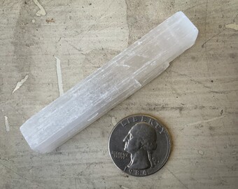 Könnte beinhalten: Ein weißer Selenit-Kristallstab, ca. 15 cm lang, neben einer US-Vierteldollarmünze. Der Kristall hat eine geschichtete Textur und ein glattes, poliertes Aussehen. Die Münze ist von 1984.