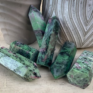 May include: Six green and pink gemstone points, also known as zultanite, arranged on a light brown surface. The points are faceted and have a natural, rough texture.