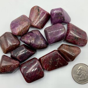 May include: A collection of ten tumbled ruby stones, each with a unique shape and a deep red color with purple hues. The stones are smooth and polished, with a glossy finish.