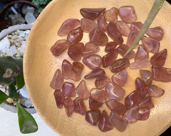 May include: A collection of tumbled rose quartz stones in various sizes and shapes, displayed on a wooden plate. The stones are a light pink color with a translucent appearance.