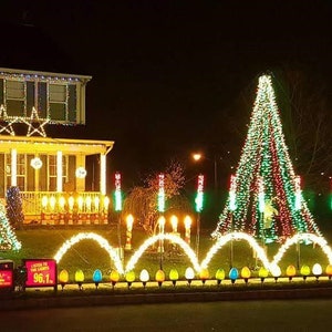 Puede incluir: Una casa decorada con luces navideñas por la noche. La casa tiene luces en forma de estrella y un árbol de Navidad hecho de luces. El jardín tiene una exhibición de luces, incluyendo una fila de bombillas grandes y coloridas.