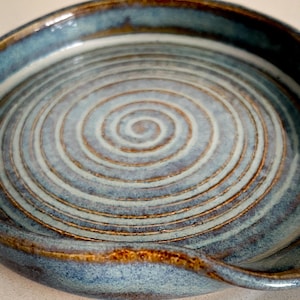 May include: A handmade ceramic spoon rest with a spiral design. The spoon rest is glazed in shades of blue and brown, with a glossy finish. The circular design is centered in the middle of the spoon rest.