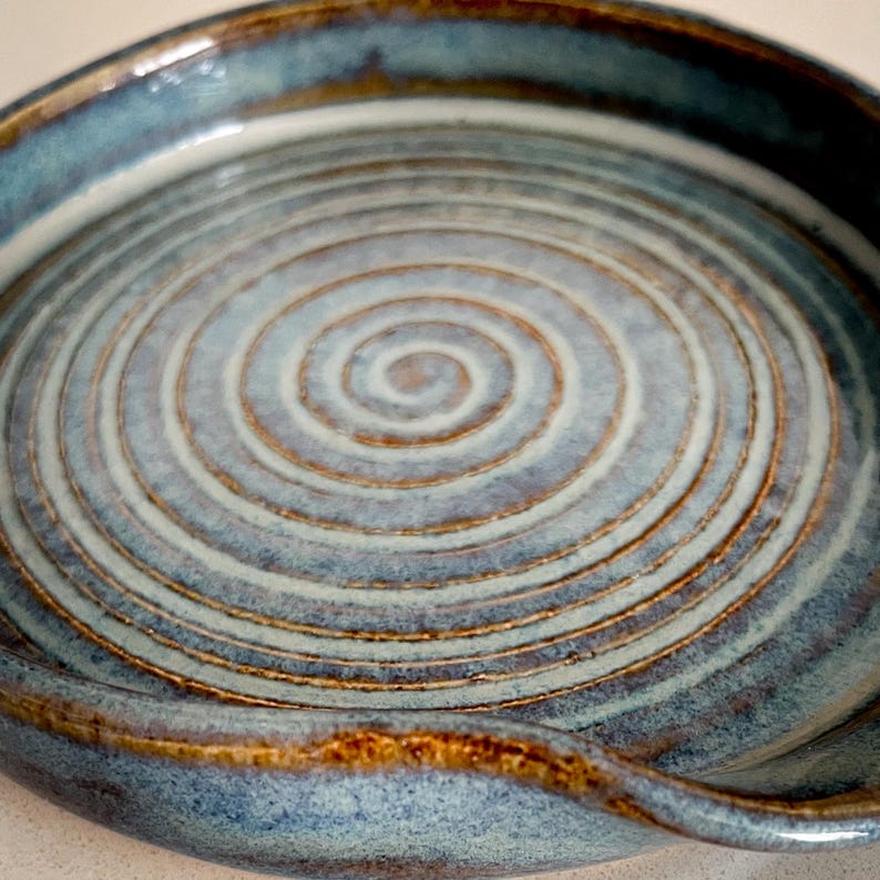 May include: A handmade ceramic spoon rest with a spiral design. The spoon rest is glazed in shades of blue and brown, with a glossy finish. The circular design is centered in the middle of the spoon rest.